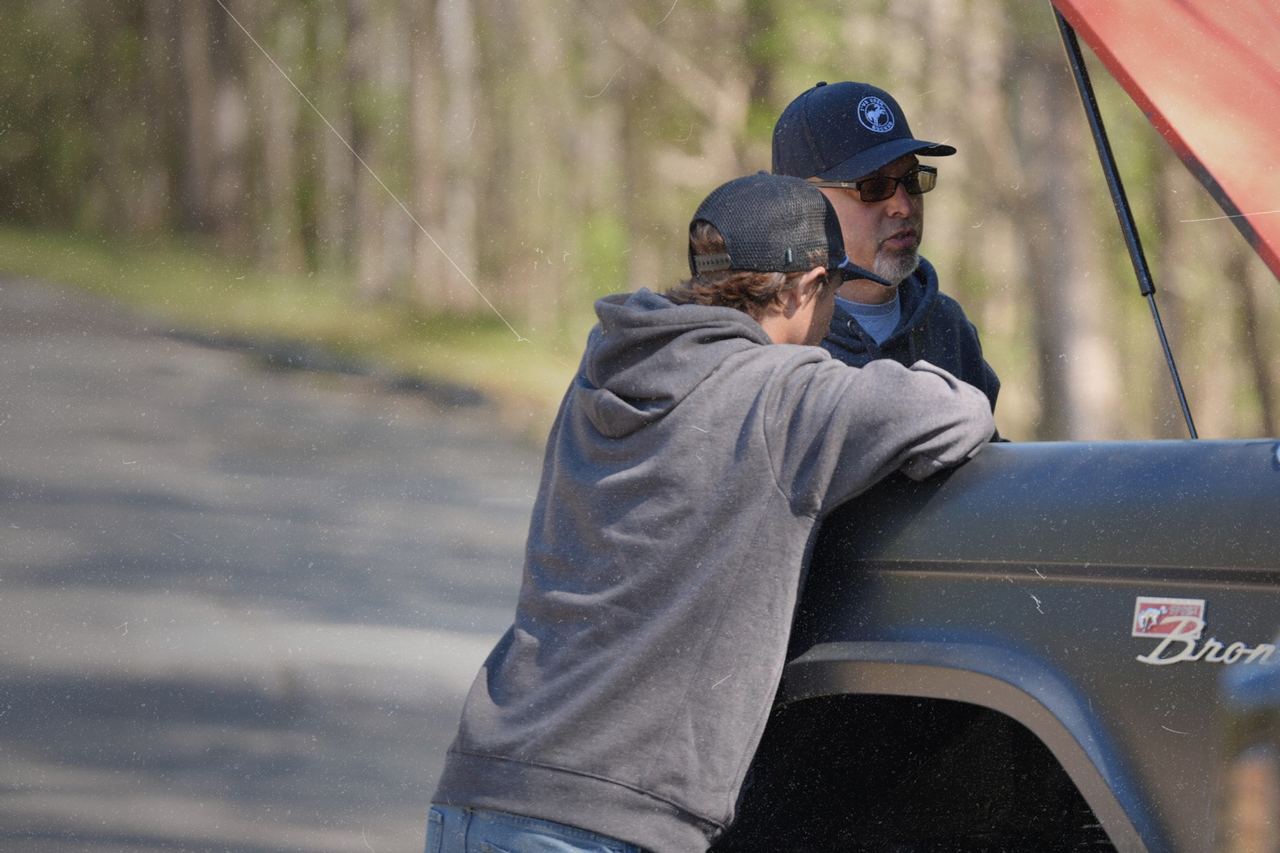 Man in IBB trucker hat drinking from a mug by a campfire, Bronco in background